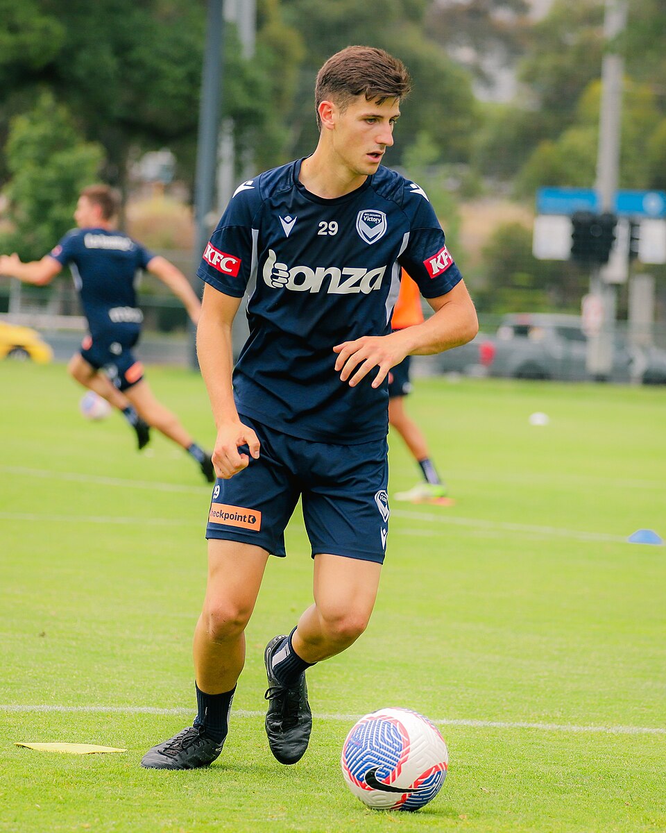 Fußballspieler im dunklen Trainingsanzug bewegt den Ball mit dem Fuß und blickt seitlich über das Feld.